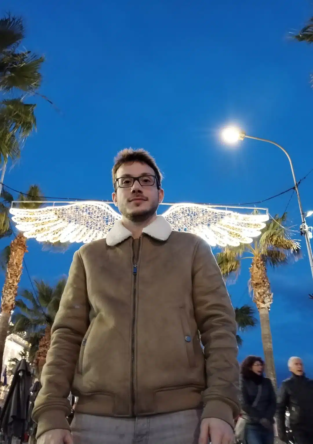 Albi - Un espace sécurisant, pour cheminer vers soi - Une personne se tient devant des ailes d'ange illuminées au crépuscule, créant un effet de halo. La rue est bordée de palmiers et des gens se promènent en arrière-plan.