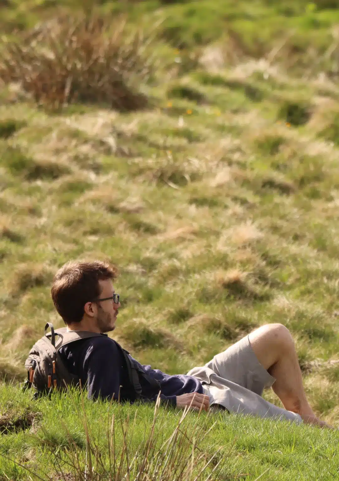 Martin Grouiller - Cadre d’accompagnement humain, securisant et empowerment - Un homme vêtu de vêtements décontractés et portant des lunettes se détend sur une colline herbeuse, adossé en arrière. Scène diurne à l'atmosphère calme et sereine. La colline est luxuriante et verdoyante.